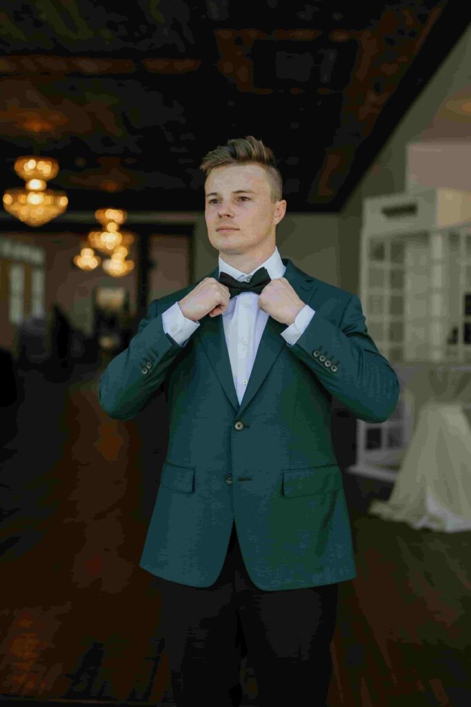 Fraiser in an olive green suit and black bow tie adjusting his jacket before the wedding ceremony.