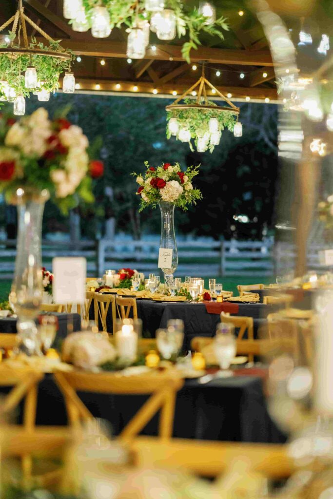 Elegant outdoor wedding reception setup with string lights, floral centerpieces of red and white flowers, and mason jar chandeliers above candlelit tables.