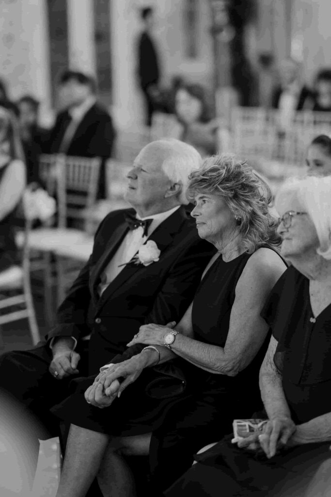 Fraiser in an olive green suit holding his bride’s hand in her white dress, with wedding guests looking at them lovingly during the ceremony.
